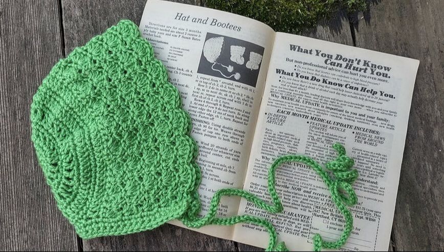 baby bonnet in green Bernat Baby Softee Yarn with a Workbasket Magazine pattern in the background